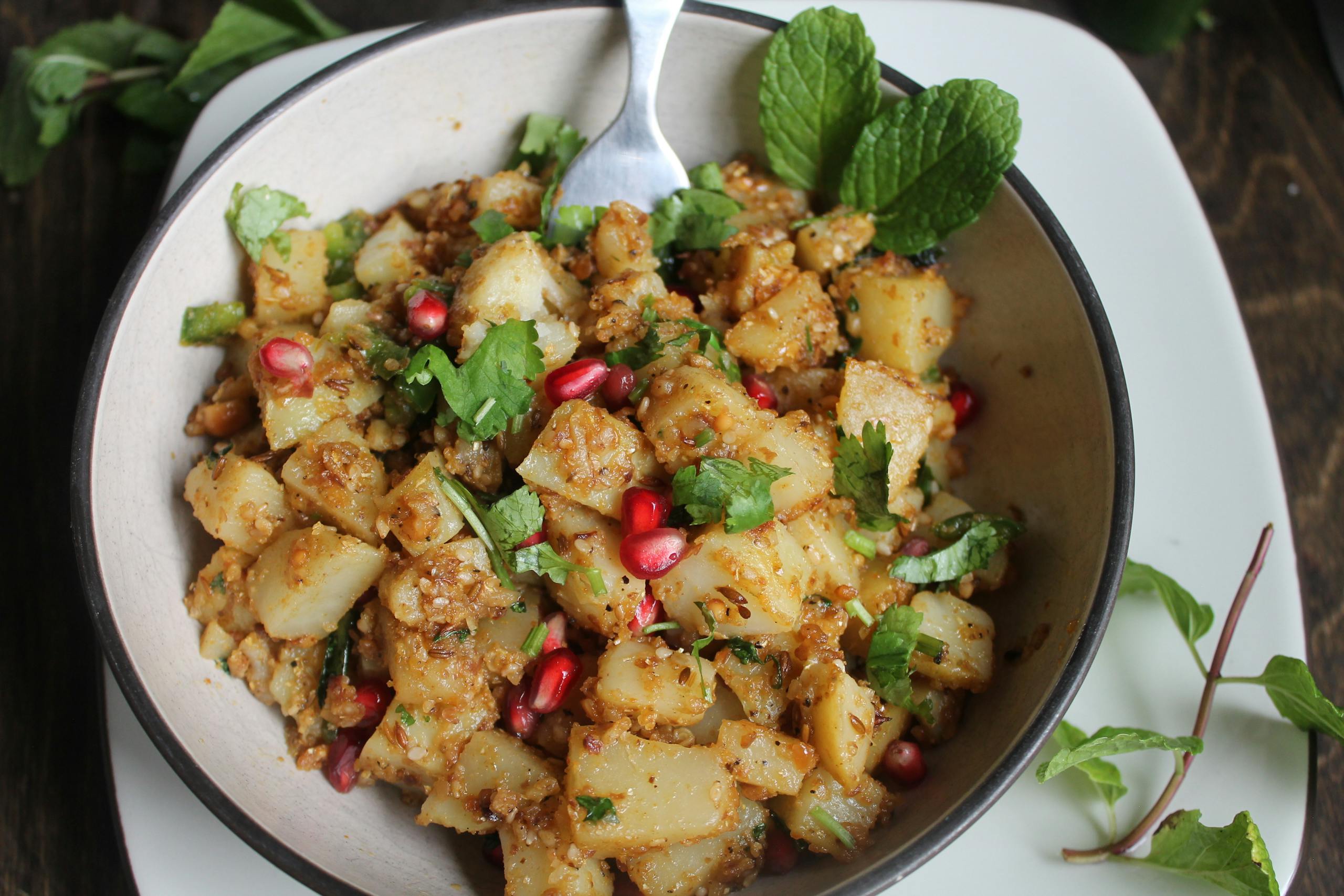 Delicious potato salad garnished with fresh mint, coriander, and pomegranate seeds in a bowl.