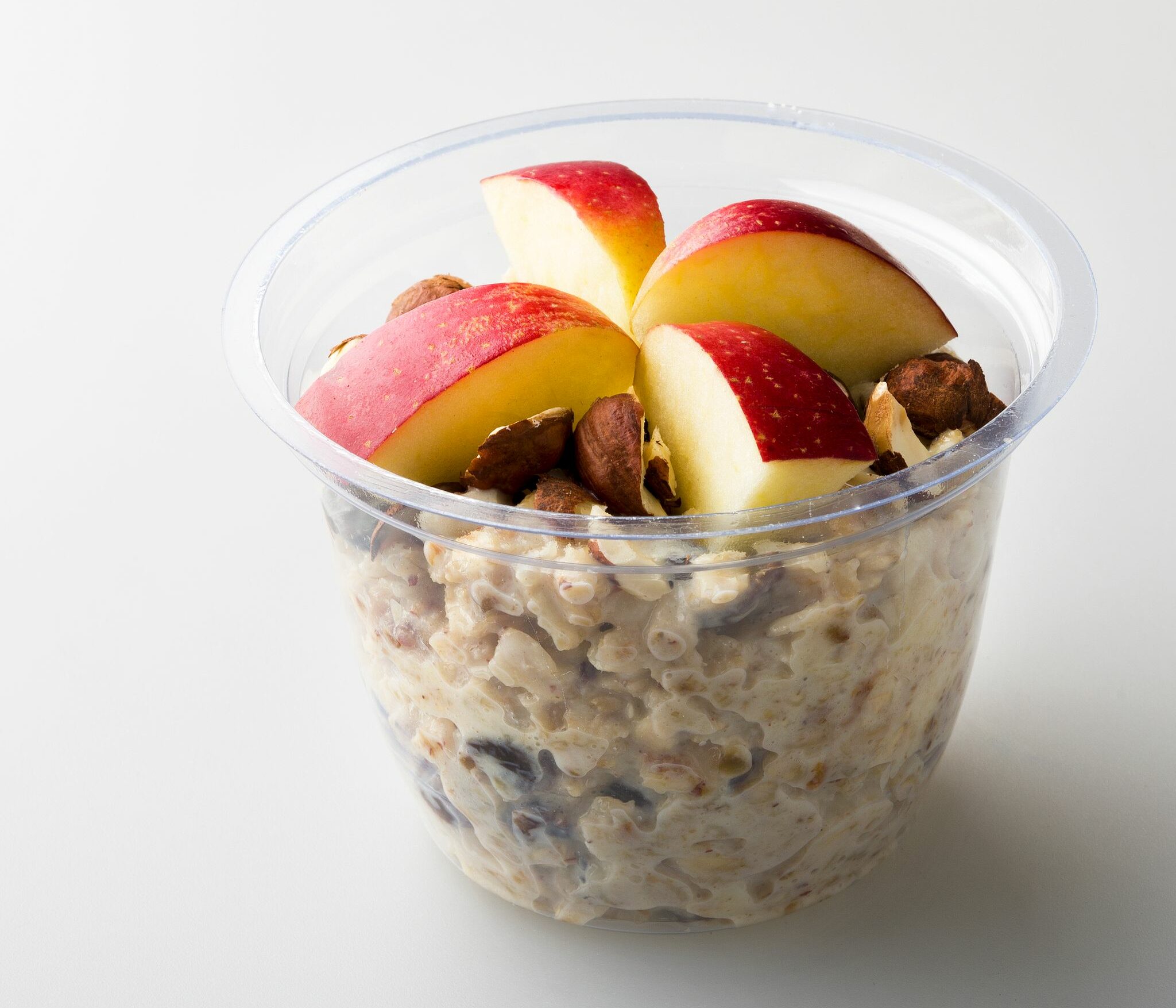 A clear plastic container of oatmeal topped with fresh apple slices and nuts on a white background.