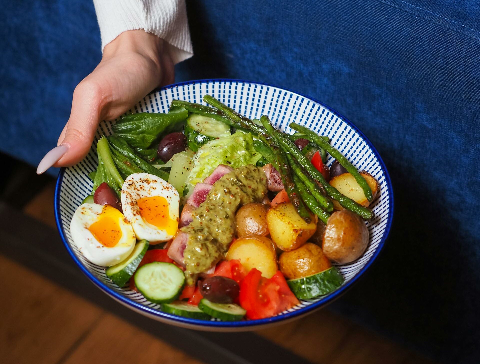 A colorful vegetable salad bowl with eggs and potatoes held by a person in a cozy setting.