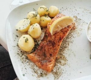 Close-up of baked salmon with potatoes, herbs, and lemon slice on a ceramic plate.