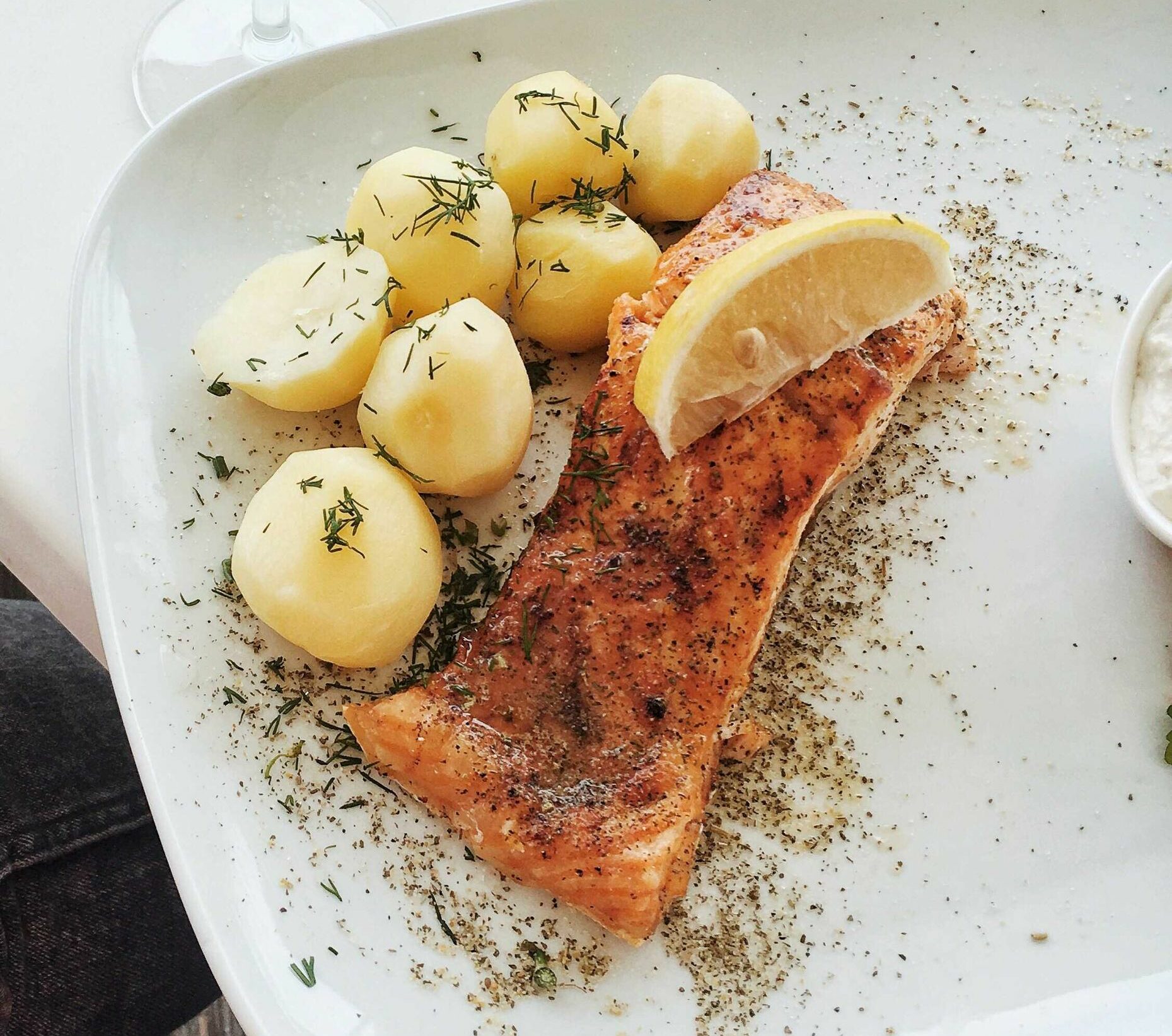 Close-up of baked salmon with potatoes, herbs, and lemon slice on a ceramic plate.