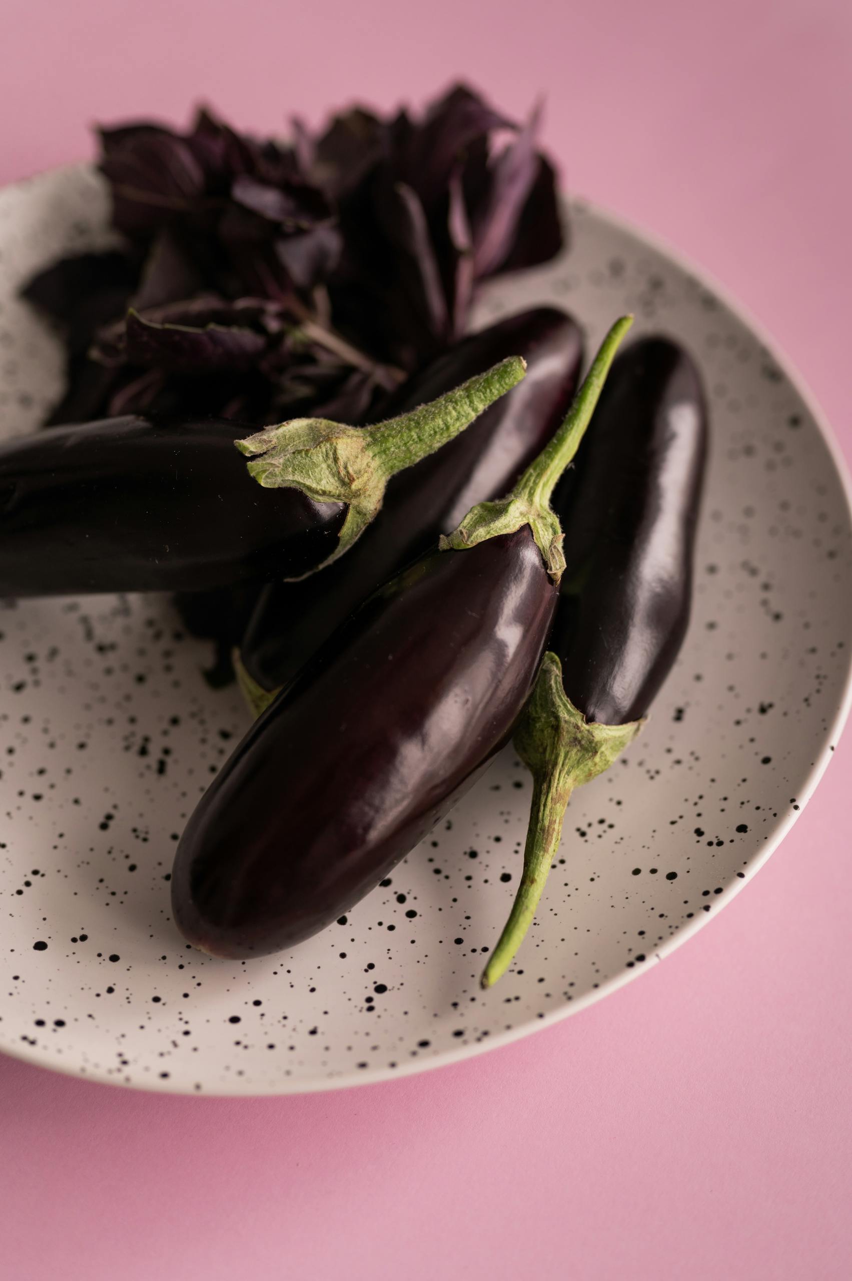 Pile of fresh ripe dark eggplants with stem and red basil placed on white plate on pink surface