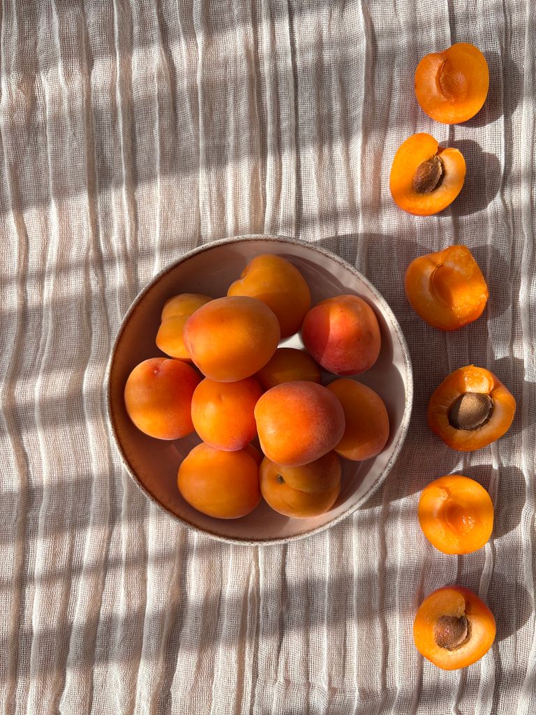 A bowl of fresh apricots with sliced halves on striped linen, lit by sunlight.