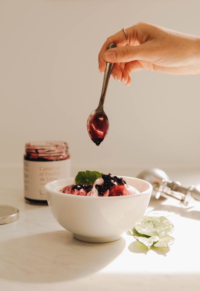 Close-up of a hand adding jam to berry ice cream in a white bowl, with natural lighting.