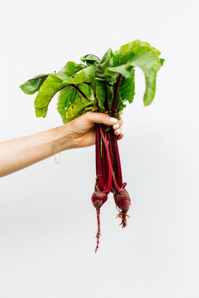 A hand holding fresh beetroots with vibrant green leaves on a white background.