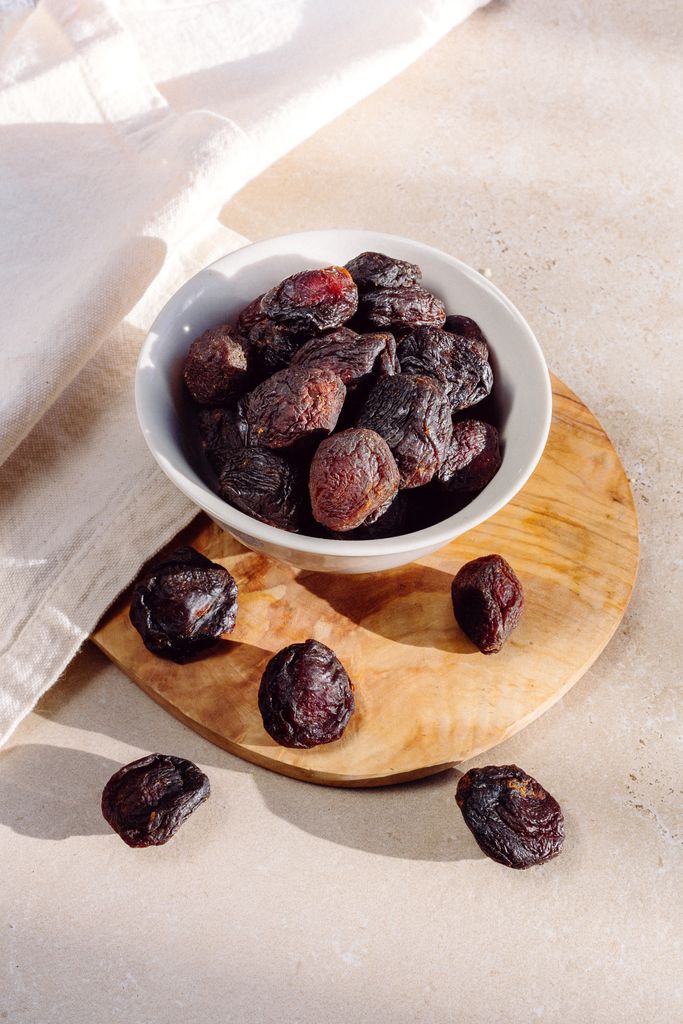 High-quality photo of dried prunes in a white bowl, set on a wooden board with natural lighting.