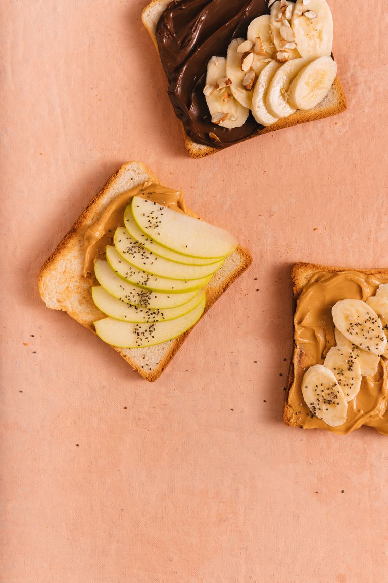 Artistic flat lay of toasts topped with nut butter, chocolate spread, apple slices, and bananas on a peach background.