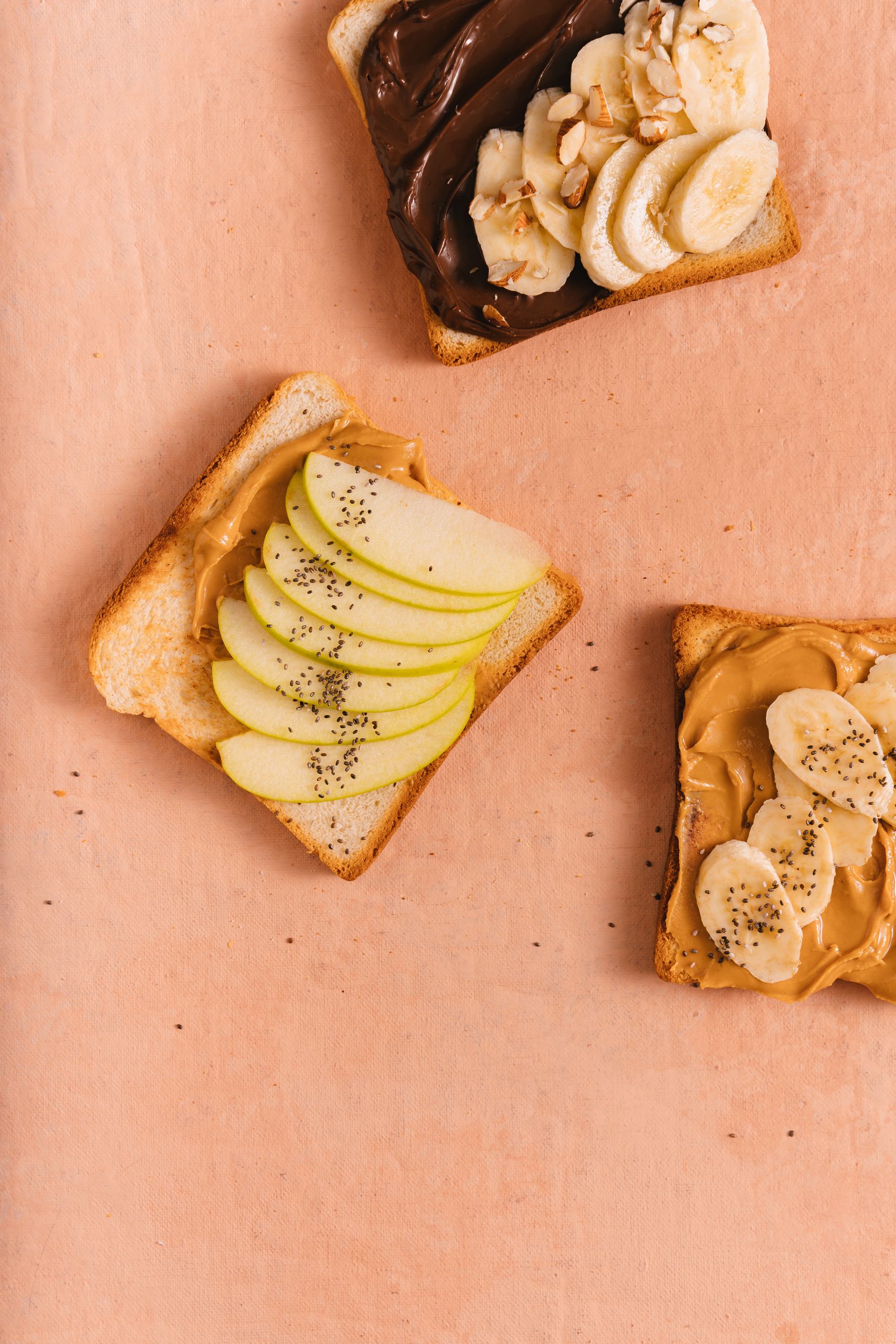 Artistic flat lay of toasts topped with nut butter, chocolate spread, apple slices, and bananas on a peach background.